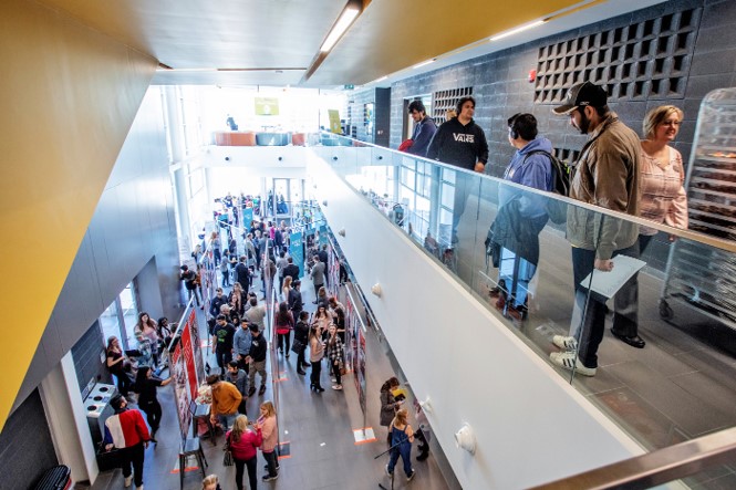 Crowds of students, staff, and visitors attending a research and innovation showcase across two levels of a campus building.