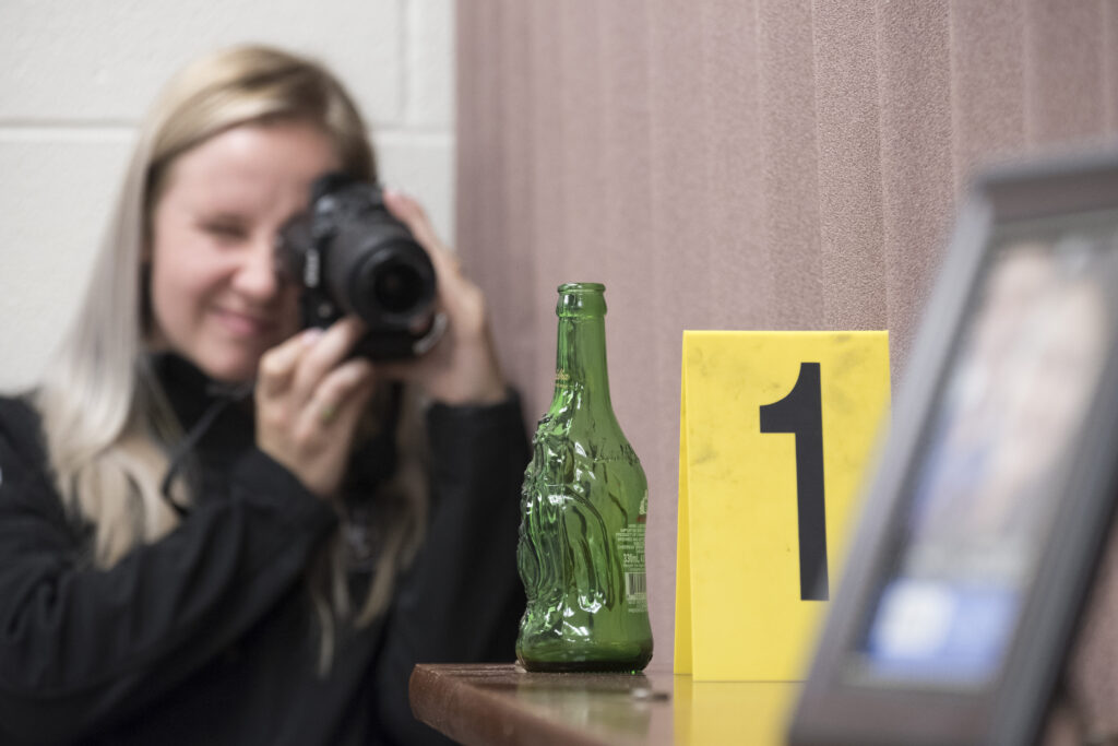 A student uses a camera to take a photo of a bottle at a crime scene.