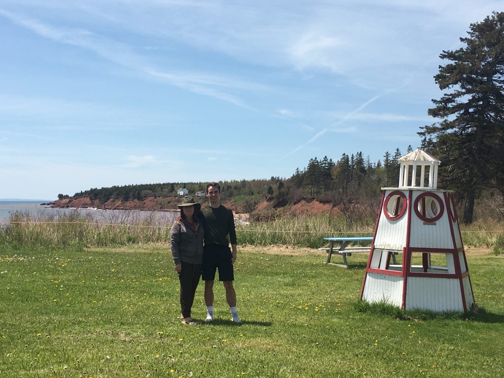 Two people smile at the camera while standing at a tree-lined lakeshore.