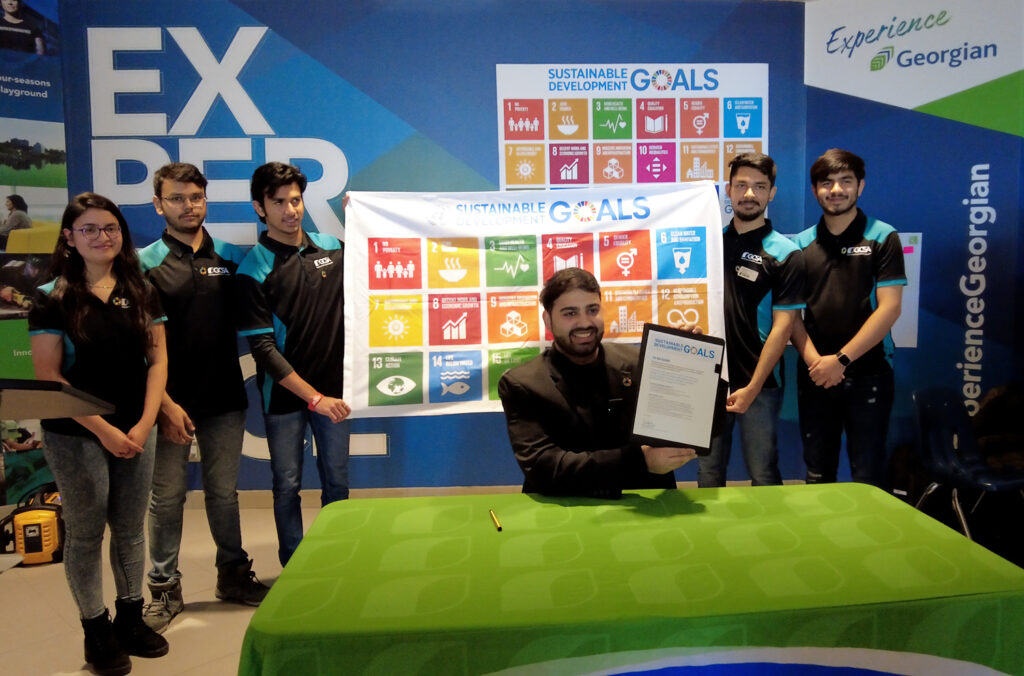 Ishaan Sachdeva, President of the Georgian College Students' Association (Barrie) holding the Sustainable Development Goals Accord, surrounded by other students and a banner of the Goals. 