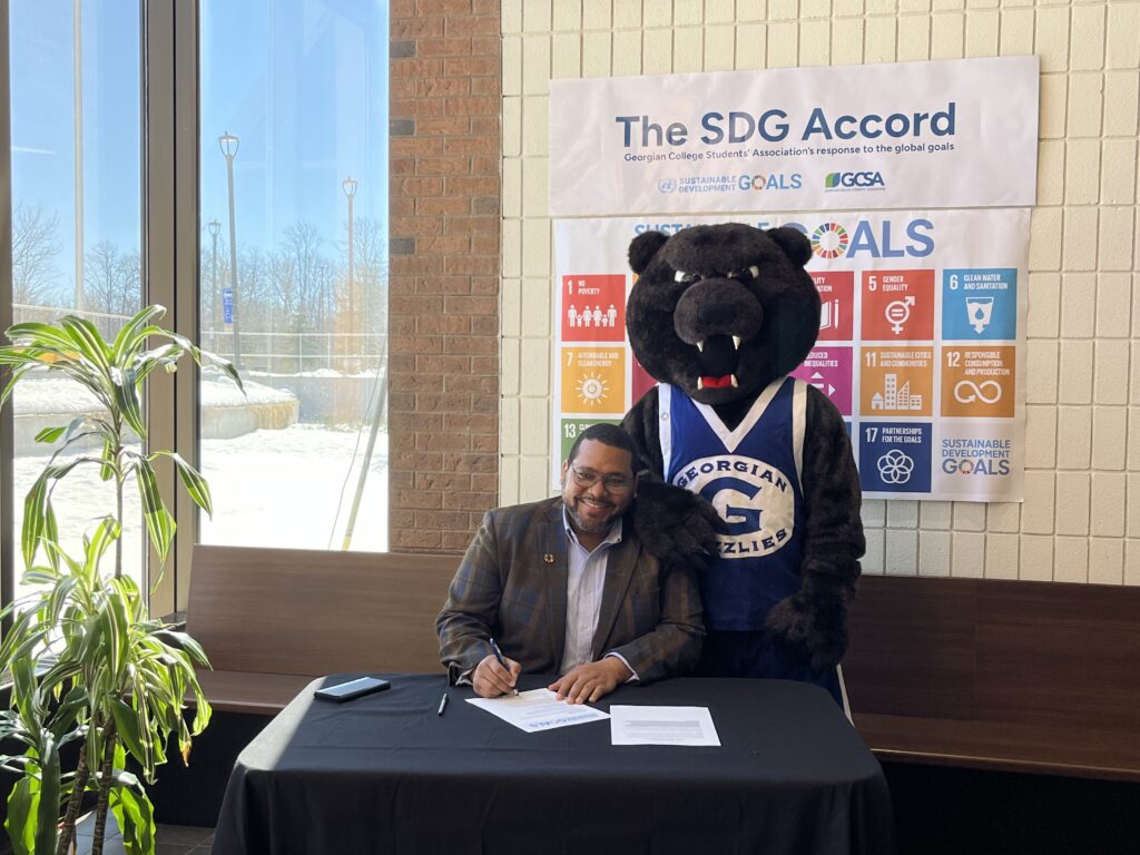 Jermaine Jones, VP Community Engagement for the Orillia Georgian College Students' Association sits with a bear mascot, signing a paper.