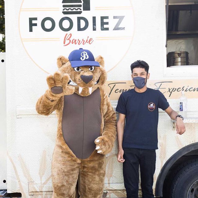 A person with short, brown hair and wearing blank pants and a navy T-shirt and face mask stands next to a cat mascot wearing a blue cap with the letter "B" on it, in front of a food truck that says Foodiez Barrie.