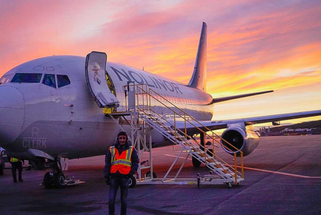 A person dressed in jeans, coat, gloves and an orange vest stands outside of a parked large plane, with its door open and stairs attached in front of a pink and yellow sunset.