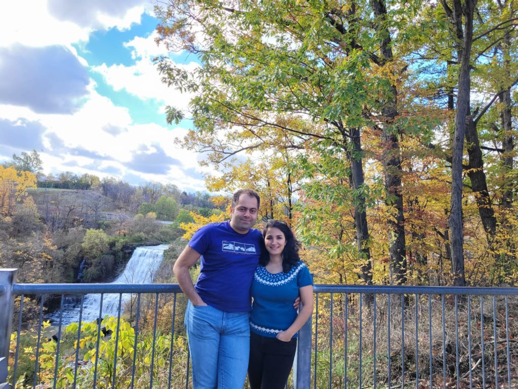 Two adults stand against a fence that blocks access to woods and a waterfall in the background.