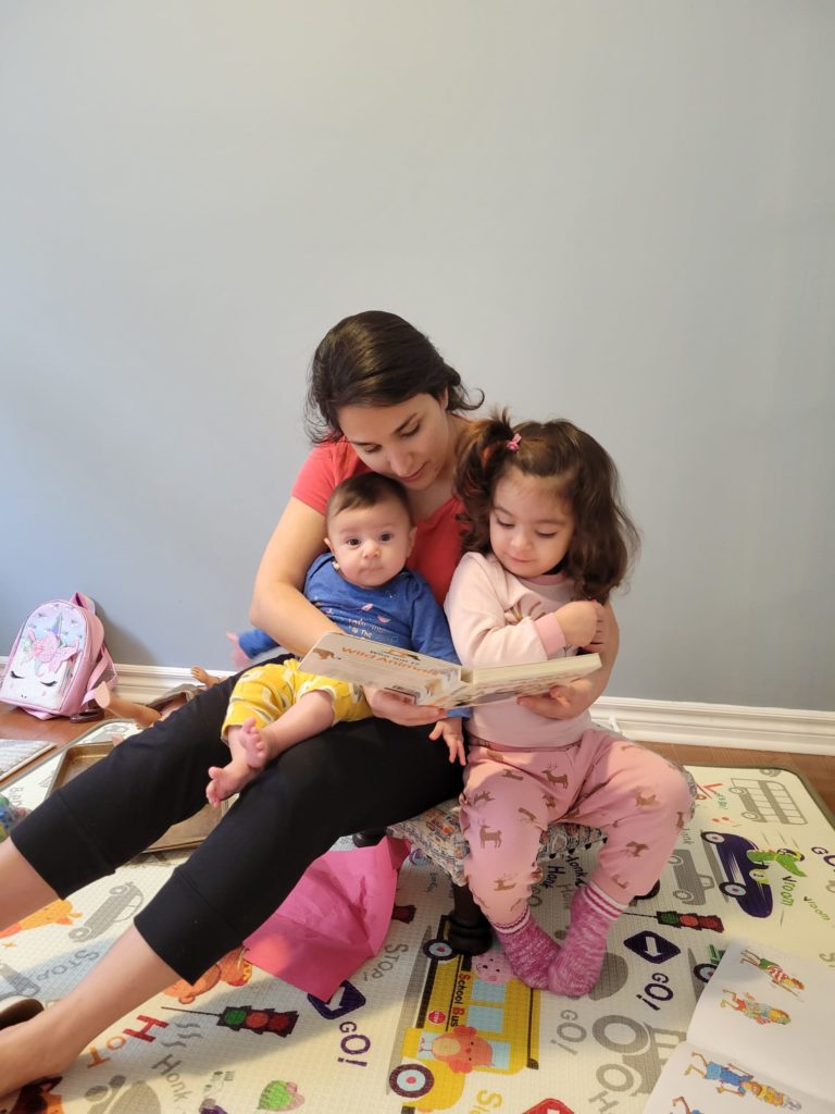 An adult sits on a stool with two children on their lap and reads a book to them. One of the kids looks up at the camera.