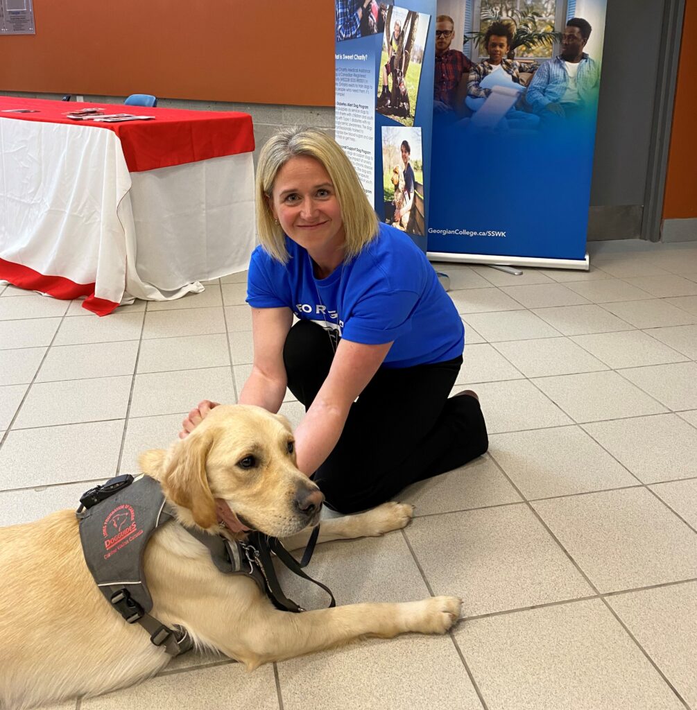 A person with blonde hair wearing a blue shirt and black pants, sitting with a Labrador therapy dog. 