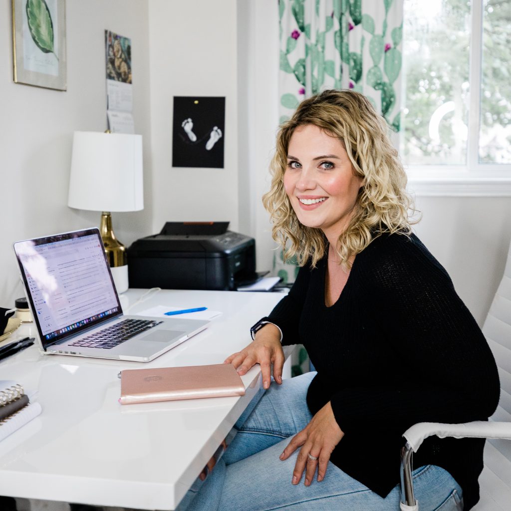 A person smiles at the camera while sitting at an office desk with a laptop and notebook on the table.