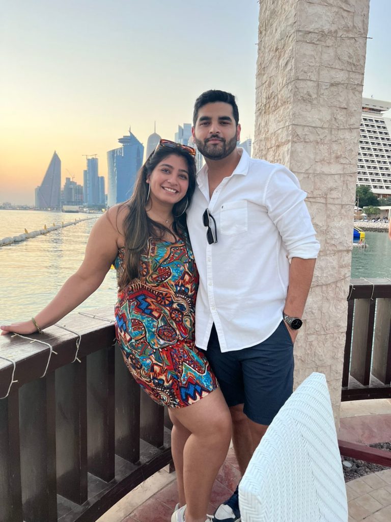 Two people stand together outside on a deck overlooking a body of water and a skyline in the distance.