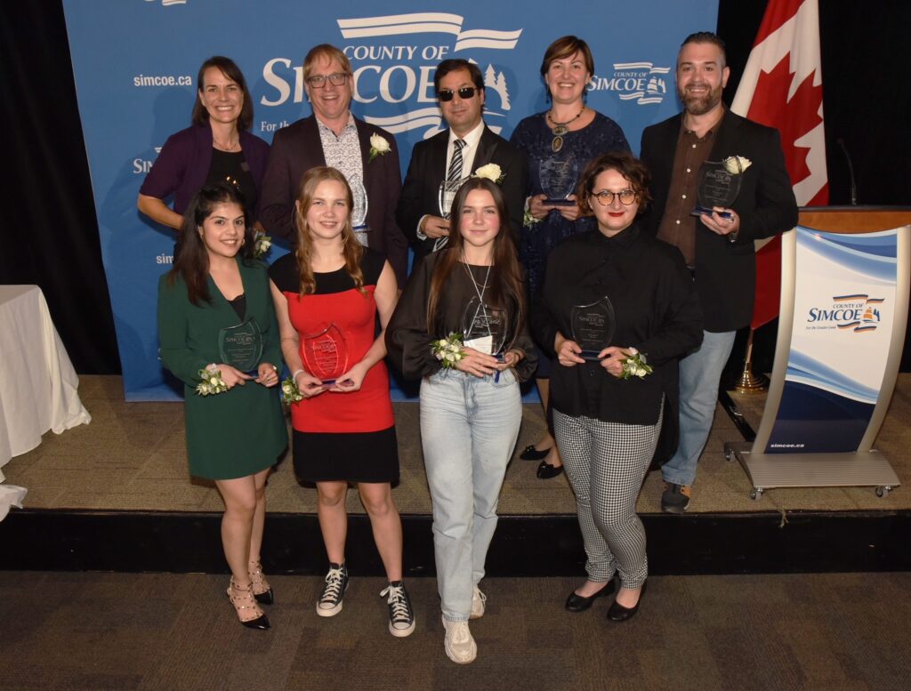 County of Simcoe Newcomer Recognition Award winners at the 11th annual event. Nine people standing on a stage with clear awards.