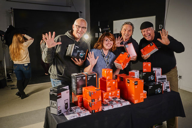 three men and one woman waving and standing in front of a table of camera equipment and boxes