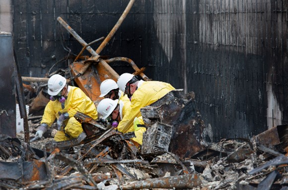 Four people wearing yellow hazmat suits and white helmets sift through building wreckage after a fire.