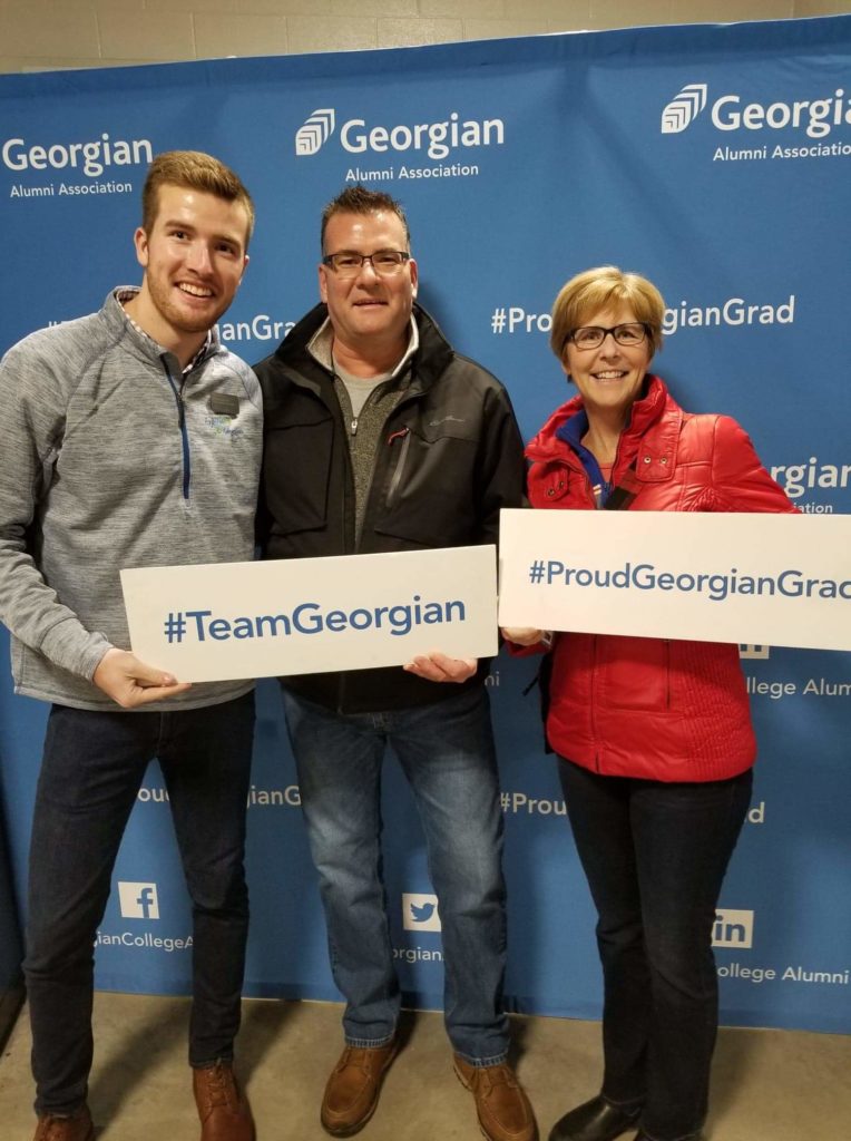 Three people dressed casually stand together in front of a blue backdrop reading #ProudGeorgianGrad. They hold signs reading #TeamGeorgian.