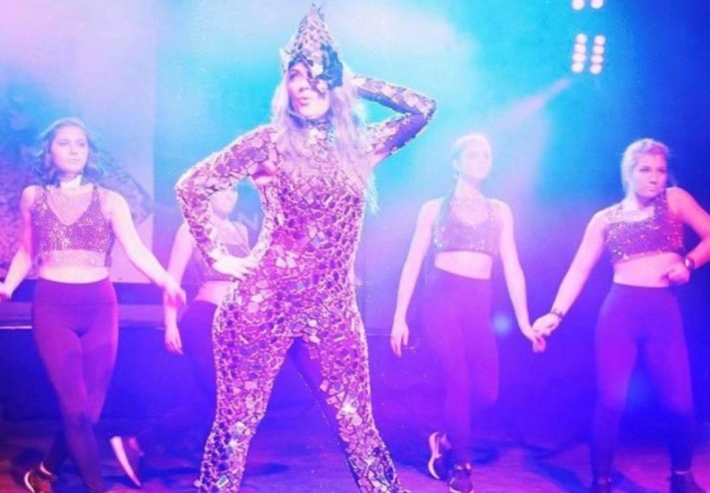 A person with long straight hair, dressed in a bodysuit covered in mirror pieces and a hat, poses on a stage with purple and blue lights around them, in front of four back-up dancers in tights and crop tops.