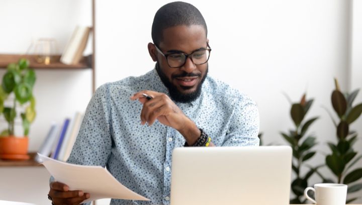 Black male with glasses wearing a blue patterned shirt, smiling while looking at this laptop.