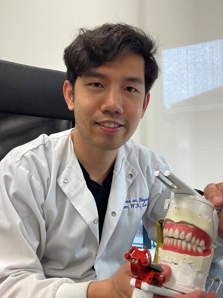 A person with short, brown hair and a white lab coat, sits in a chair and holds a model of dentures.