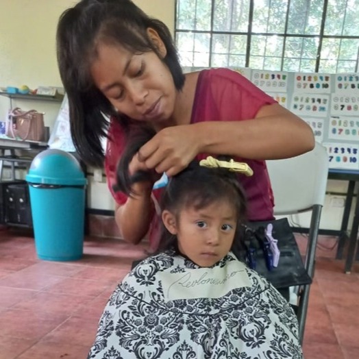 A Guatemalan woman with chin length dark brown hair cuts the hair of her toddler in the Hair Aid clinic in 2024. 
