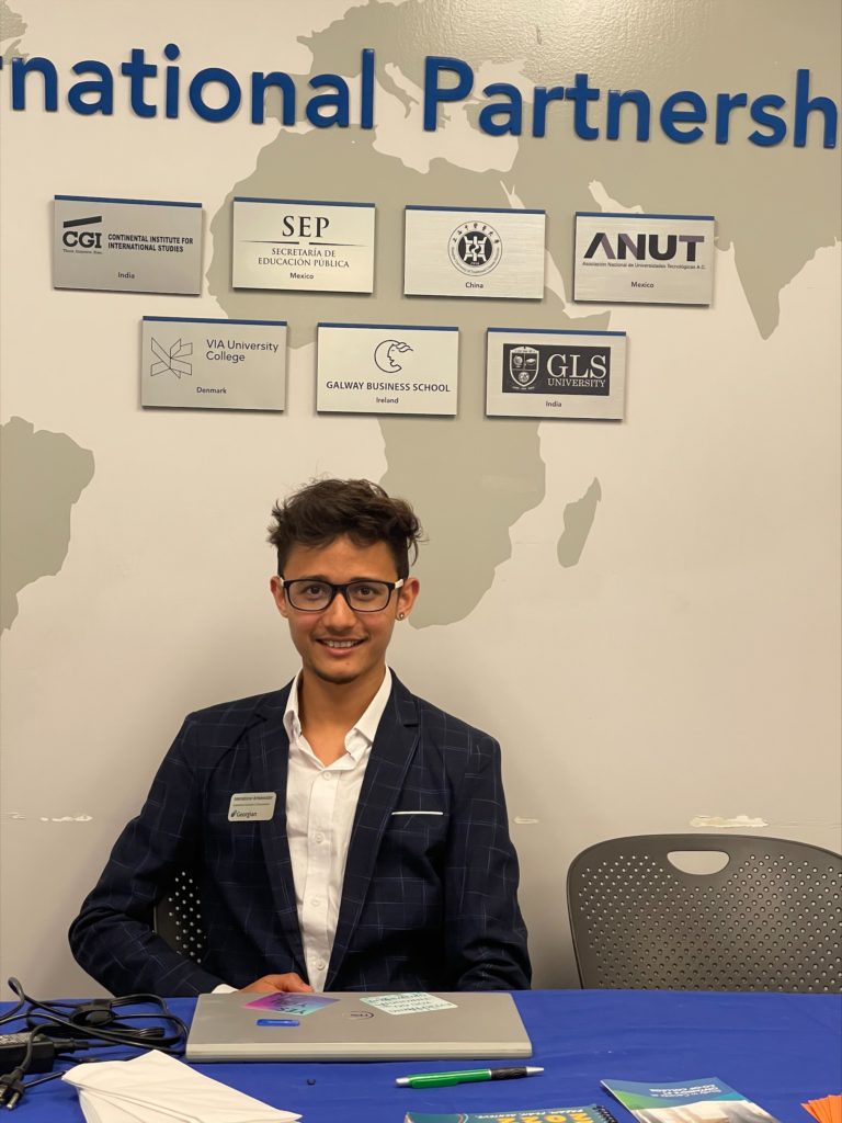 A person wearing glasses, a dress shirt and blazer sits at a table with a sign overhead reading "International Partnership."