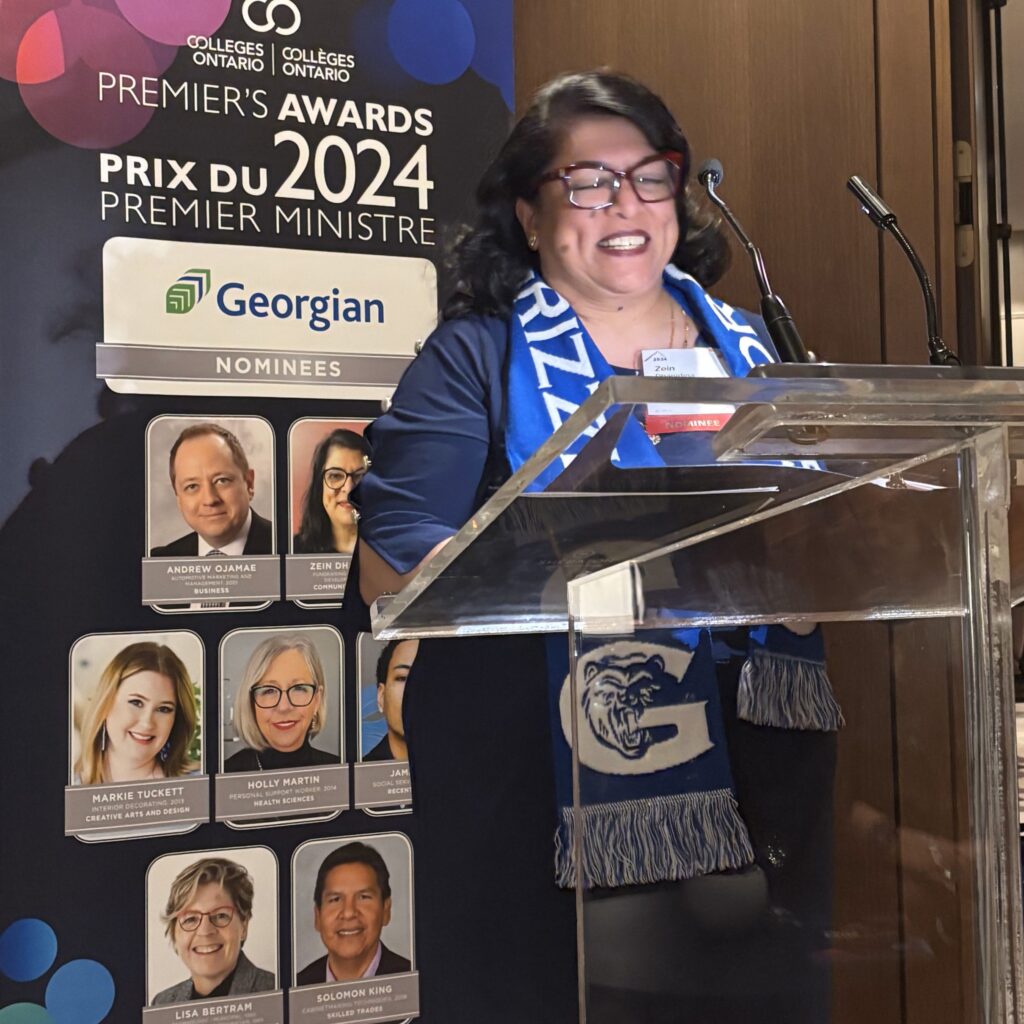 Zein Dhanidina standing at a podium at the Premier's Awards 2024 ceremony. She's wearing her dark brown hair at should length and has a Georgian Grizzlies scarf around her neck.