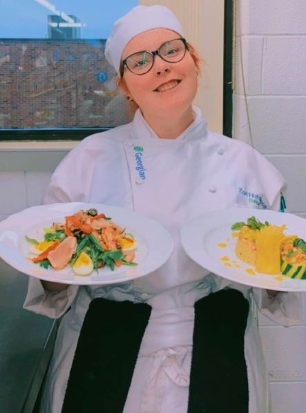 Person smiles at the camera while holding two plates of food.