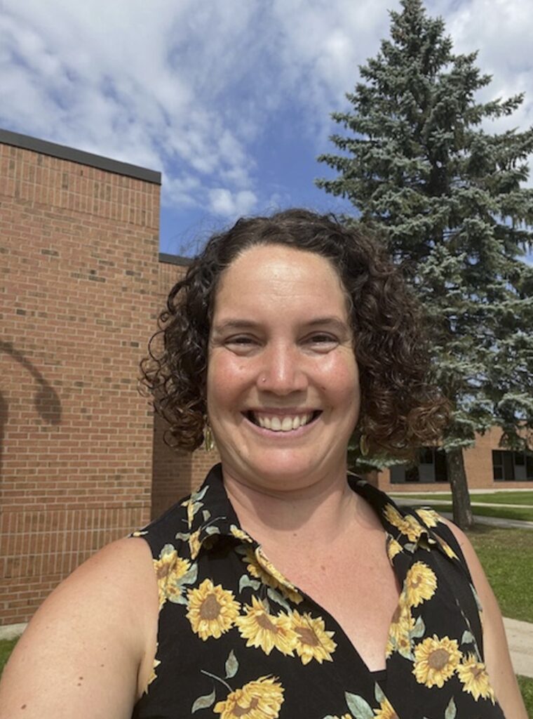A person with short, curly hair and a sunflower shirt takes a selfie outside in front of buildings and a tree. 