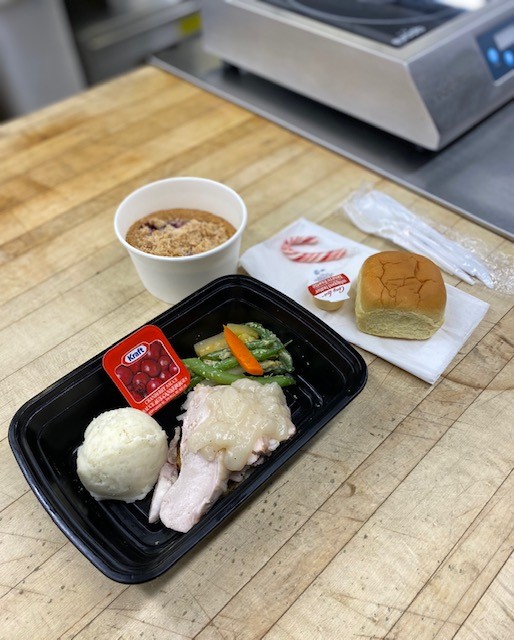 A container with potatoes, turkey, dressing and vegetables on a wooden counter. There is a container with dessert beside it and a bun and a candy cane on a napkin.