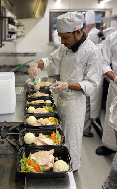 A young male wearing a white chef uniform putting food into black containers in a kitchen