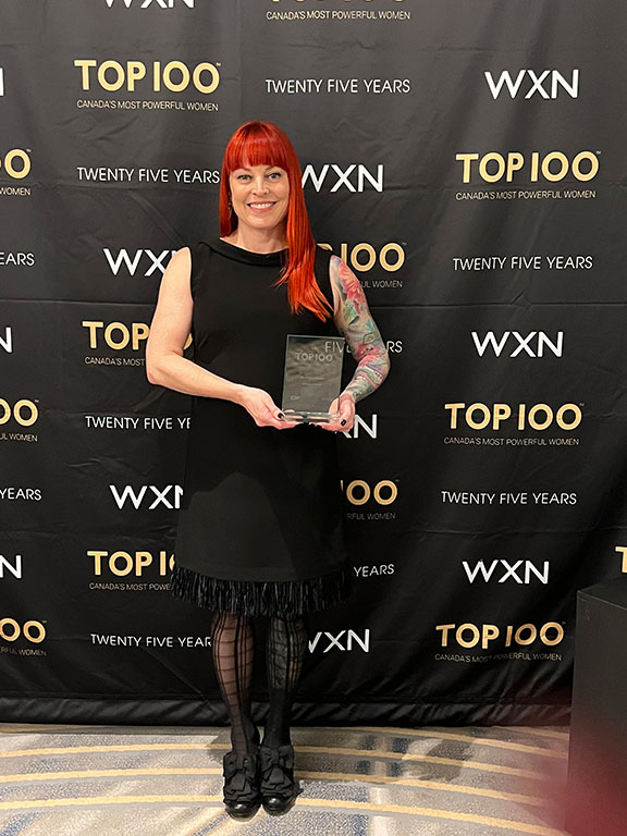 A smiling female (Brandi Ferenc) with long red hair wearing a black dress. She's holding a glass award and is standing in front of a backdrop that says Top 100