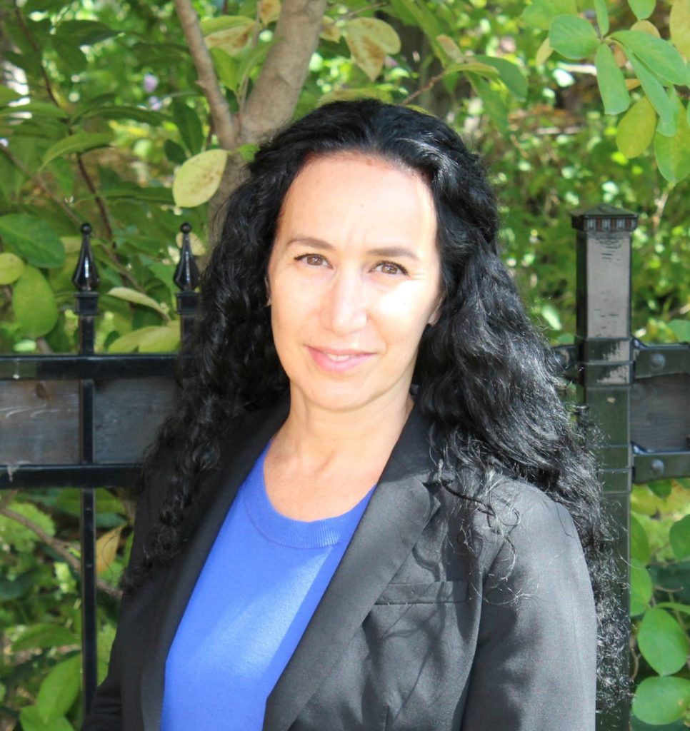 A smiling white woman with long black hair wearing a blue shirt and a grey jacket. She's standing outside.