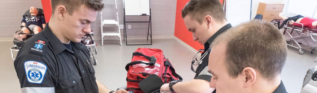 Three Advanced Care Paramedic students in uniform organizing equipment in red first-aid kits in a simulation lab