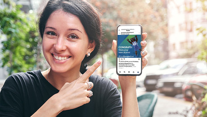 Person with dark hair pinned back in a pony tail standing outside while holding and pointing to a cellphone