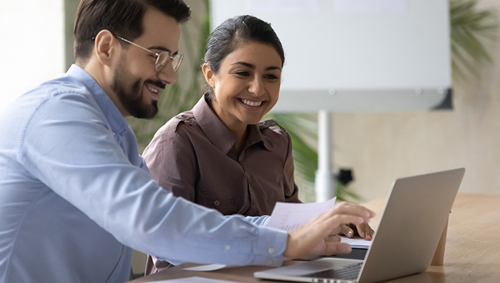 A mentor and mentee sit next to each other at a desk with a laptop and paperwork while smiling