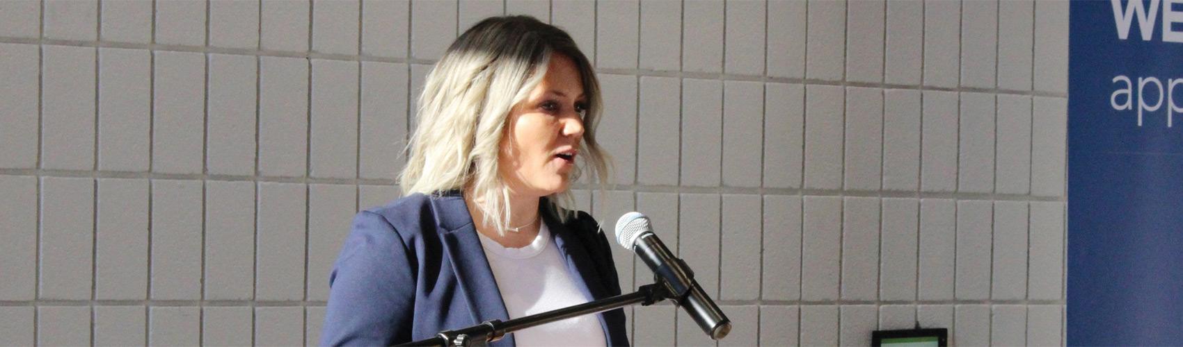 A person in a white t-shirt, navy blue blazer and with short, curly blonde hair speaking into a microphone at a podium