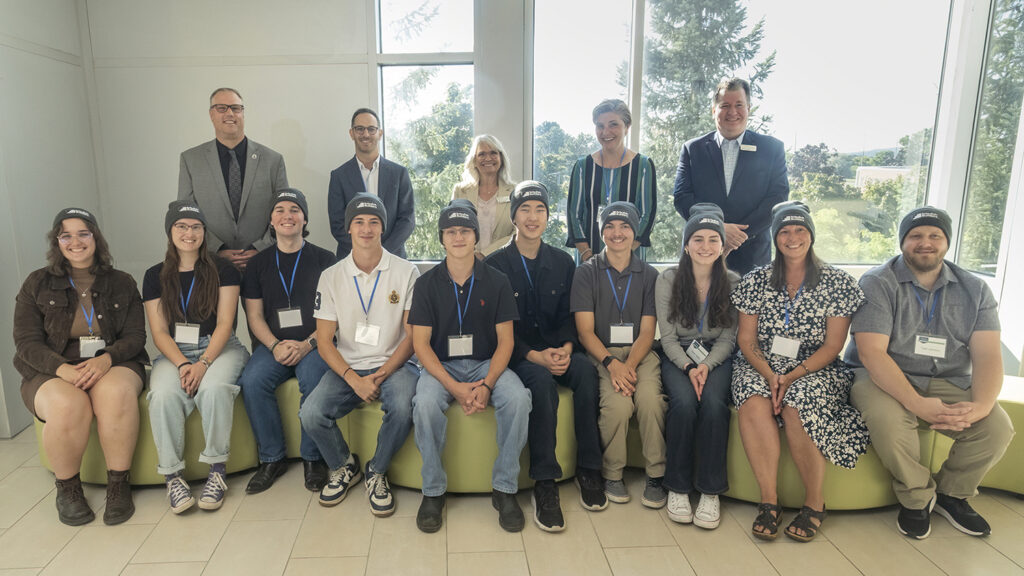 Ten people each wearing the same toque sit in a row on green couches, with five people standing behind them. 