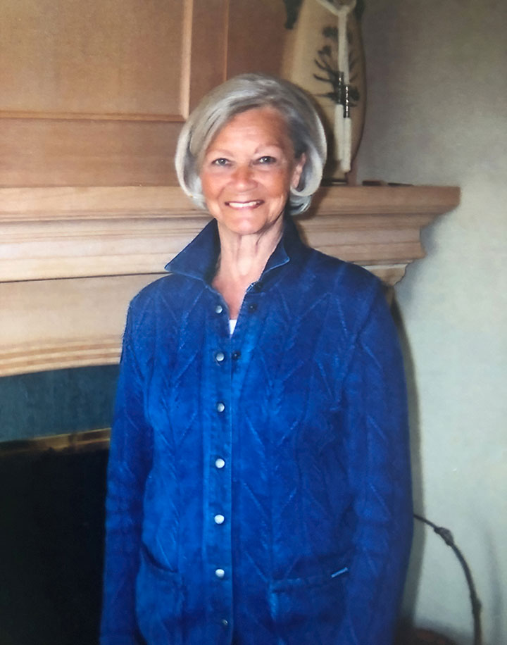 A smiling older woman with short grey hair (Verdi Wallwin) standing in front of a fireplace. She's wearing a blue shirt with long sleeves.