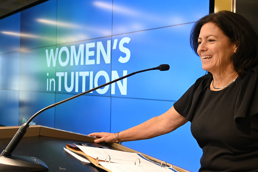 A smiling woman with short dark hair wearing a black dress (Gisele Bodkin) standing at a podium in front of a screen saying WOMEN'S inTUITION