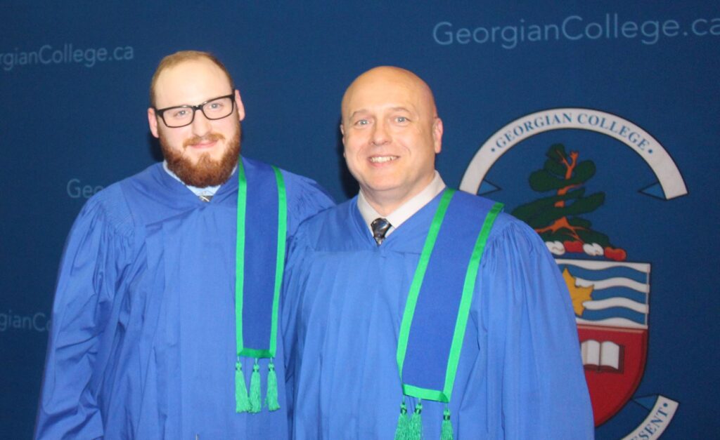 Two people wearing blue convocation robes stand next to each other in front of a backdrop reading "СŶƵ.
