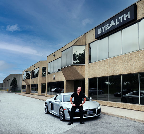 a man (Ed Boutilier) standing in front of a silver car, dressed all in black. He's outside a building with a large sign reading "STEALTH"