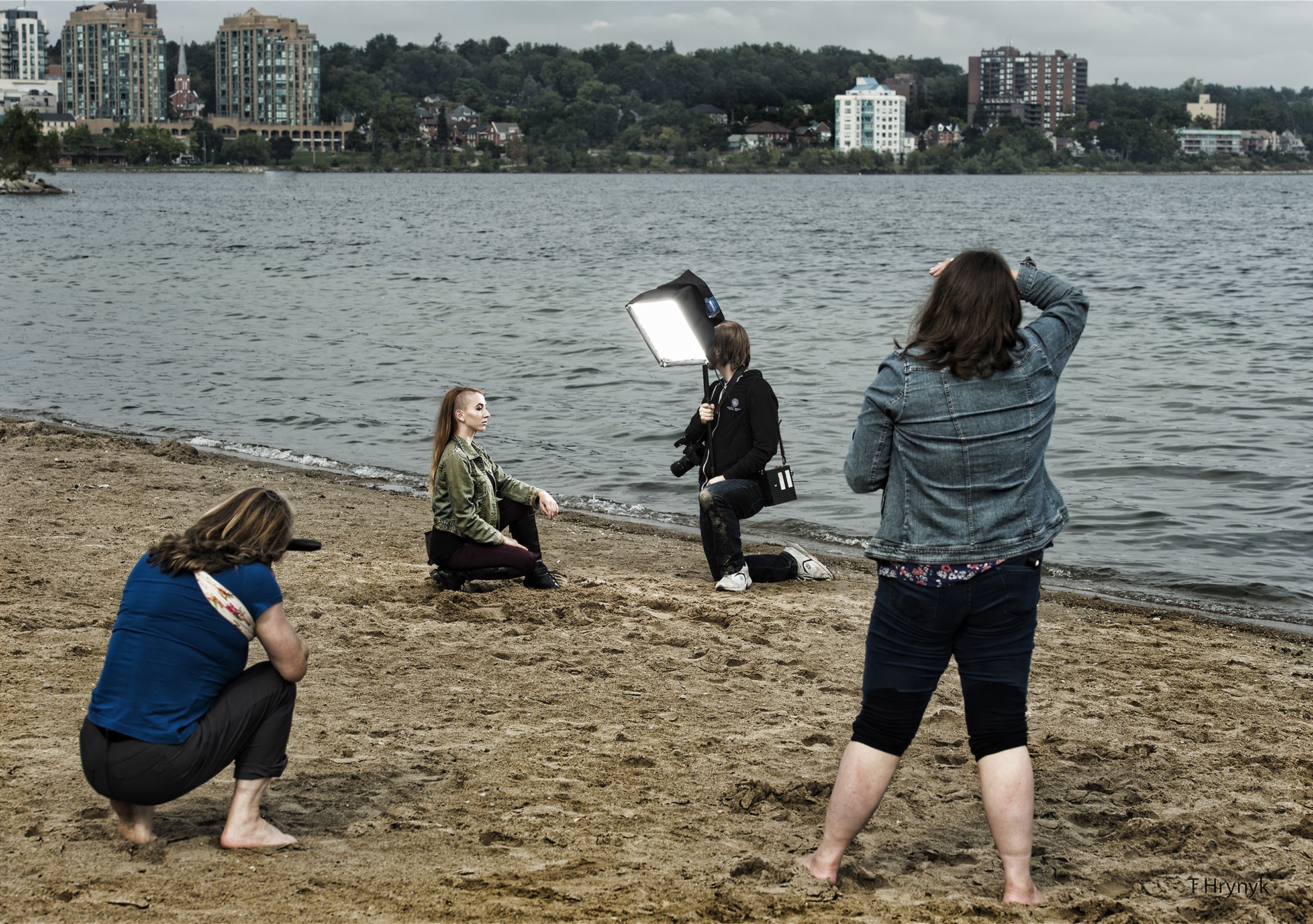 A female kneeling and posing on a beach in front of two female photographers and another female holding softbox lighting equipment