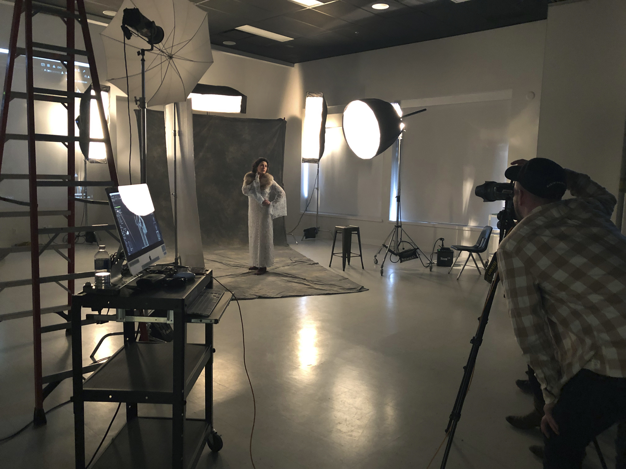 a female student dressed in a while gown posing with her arm on her hip in front of a grey backdrop while a male student captures her photograph
