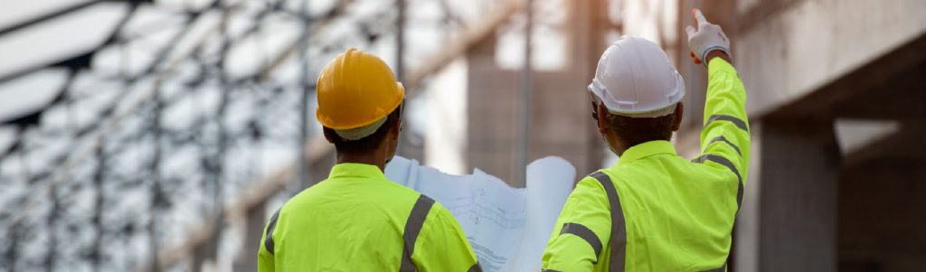Two architectural technicians in reflective yellow work coats and construction hard hats holding a blueprint and pointing to a structure
