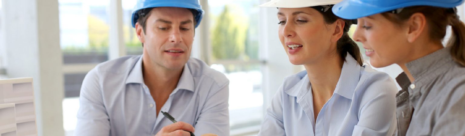 Three architectural technologists wearing dress shirts and construction hard hats while looking down at a tablet screen