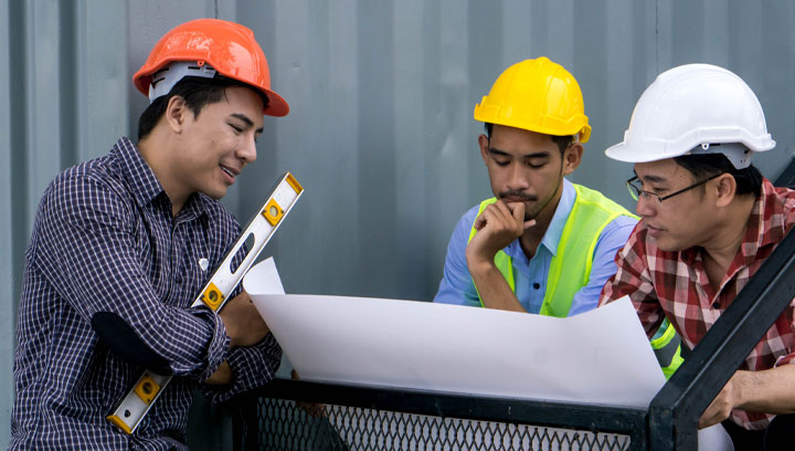 Three architectural technologists wearing construction hard hats and reviewing blueprint paperwork together