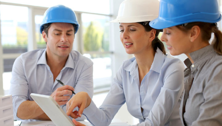 Three architectural technologists wearing dress shirts and construction hard hats while looking down at a tablet screen