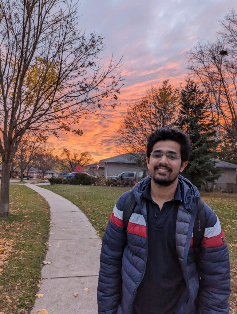 A person with short, black hair, glasses and a goatee stands outside on a sidewalk and smiles, with an orange sunset sky above.