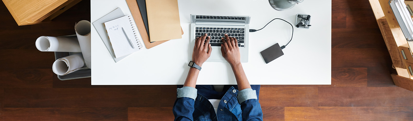 assessment banner, overview of young businessman in denim jacket sitting by desk working from laptop