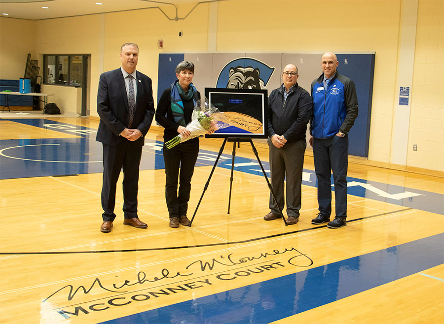 Three men (Kevin Weaver, Brian Muscat and David Laliberte) standing in a gym with Michele McConney. She's holding flowers and there's wording on the floor in front of them saying McConney Court.