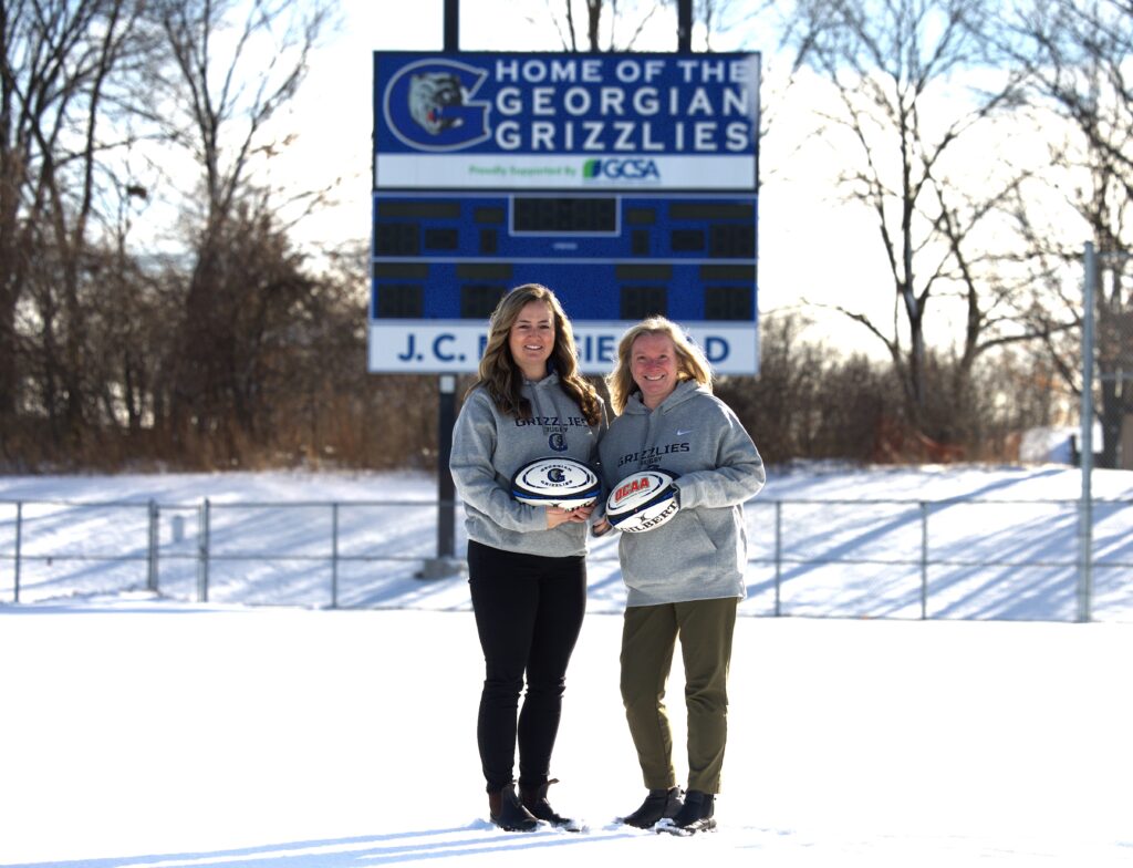 Two people stand on a snowy rugby field holding rugby balls.