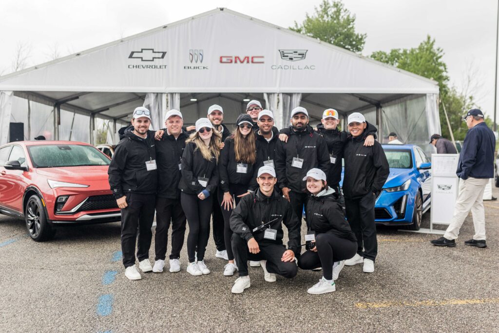 A group of people wearing matching black pants and jackets and white ballcaps stand outside in front of vehicles on display under a tent.