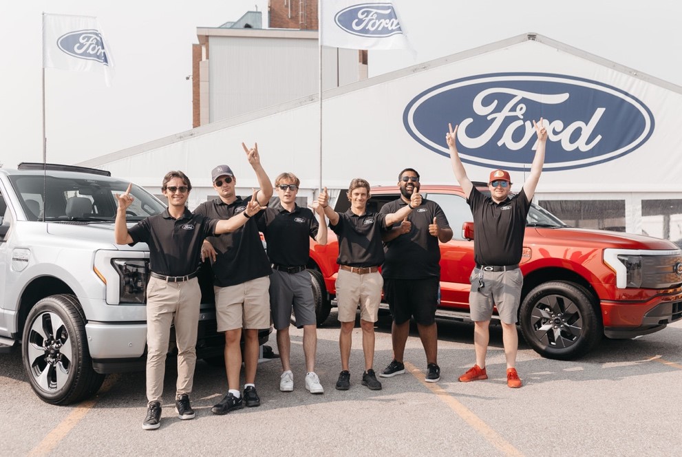 Six people wearing matching black shirts stand in front of a tent that reads "Ford" and two vehicles.
