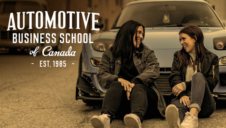 Two Automotive Business students smiling at each other while sitting on the pavement in front of a sky blue Mazda sports car